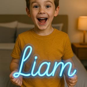 Puede incluir: Un letrero de ne&oacute;n azul con el nombre "Liam" en escritura cursiva. El letrero lo sostiene un ni&ntilde;o que lleva una camiseta de color amarillo mostaza y sonr&iacute;e. El fondo es un dormitorio.