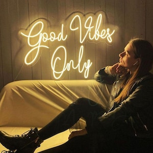 以下が含まれることがあります： 暗い木製の背景に白いネオン文字で「Good Vibes Only」と書かれたネオンサイン。白いソファに座っている人がサインの前にいる。