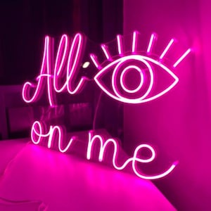 May include: Pink neon sign with the words "All on me" and a stylized eye with eyelashes.