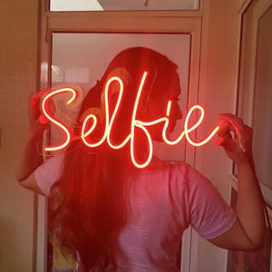 May include: A red neon sign that says "Selfie" held up by a person with long red hair.