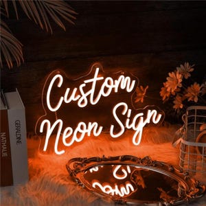 Könnte beinhalten: Ein weißes Neon-Schild mit dem Text "Custom Neon Sign" in einer kursivschriftlichen Schriftart. Das Schild leuchtet und wirft einen orangefarbenen Schein auf die Umgebung.