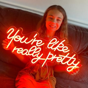 May include: A neon sign with the phrase "You're like really pretty" in a cursive font. The sign is lit with a bright, warm red glow. The sign is held by a person.