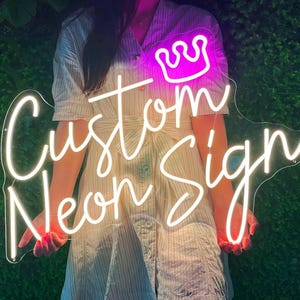 May include: A white neon sign with the text "Custom Neon Sign" in a cursive font. A pink neon crown is above the text. The sign is held by a person.