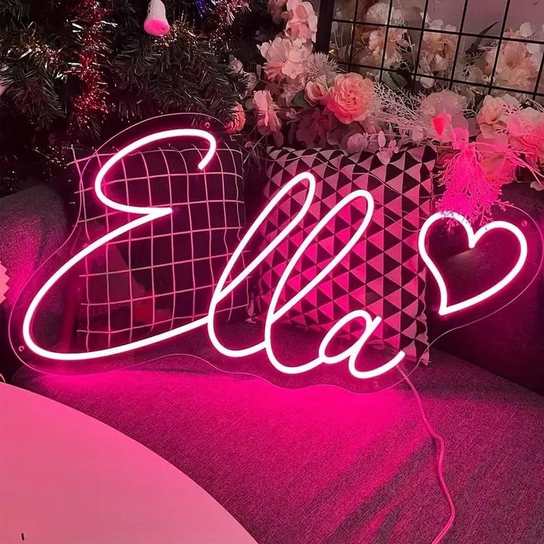 Puede incluir: Un letrero de ne&oacute;n rosa que dice "Ella" en cursiva, con un coraz&oacute;n. El letrero brilla intensamente. El fondo presenta cojines decorativos y flores, creando una escena decorativa.