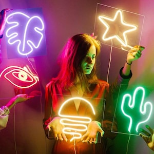 May include: A woman holds up a neon sign shaped like a star. Other neon signs are visible in the background, including a cactus, a leaf, a burger, and an eye.