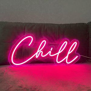 May include: Pink neon sign with the word "Chill" in a cursive font. The sign is mounted on a wall above a gray couch.