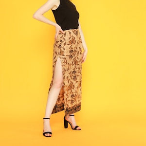 May include: A woman wearing a black sleeveless mock neck top and a long brown and black floral skirt with a slit up the side. She is wearing black heeled sandals and is standing on a yellow background.