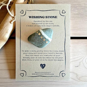 May include: A Wishing Stone on a tan card with a heart design. The stone is gray-green with white and gold accents. Text on the card reads "Smoothed by the sea and kissed by the tide, a token of luck with magic inside."