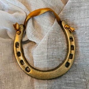 May include: A gold-colored horseshoe with a yellow ribbon tied to the top. The horseshoe has seven holes and is resting on a textured, light-colored fabric. The ribbon is looped and tied in a knot.