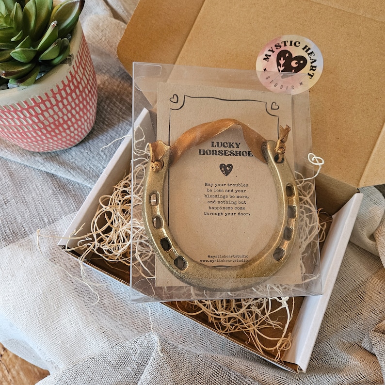 May include: A gold-colored horseshoe with a brown ribbon, presented in a clear box with a card that reads "Lucky Horseshoe." The card includes a message about blessings and happiness. The box is inside a cardboard box.