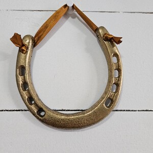 May include: A gold-colored horseshoe ornament with a bronze ribbon for hanging. The horseshoe has a textured surface and is suspended against a white background. The horseshoe is a symbol of good luck.