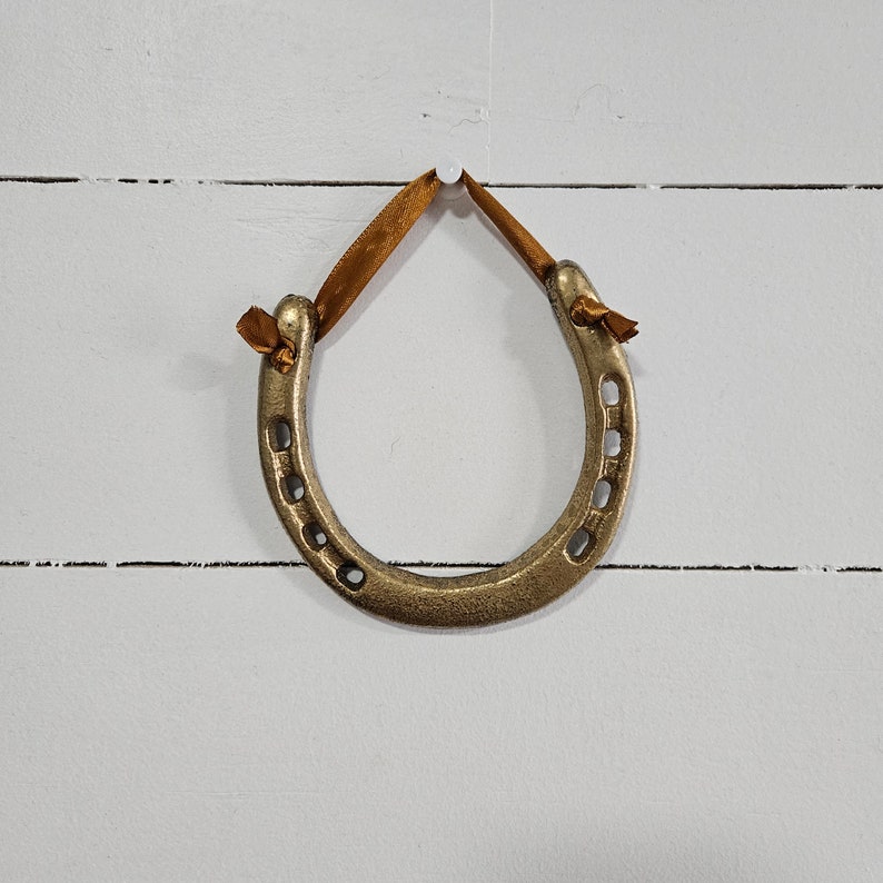 May include: A bronze-colored horseshoe ornament with a brown ribbon for hanging. The horseshoe has a classic U-shape with seven holes and is suspended against a white, horizontal-slat background.