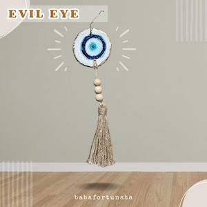 May include: A hanging wall ornament featuring a circular white disc painted with a blue and white evil eye symbol. The disc is suspended by a string with three light-colored wooden beads and a long, textured tan jute tassel at the bottom.
