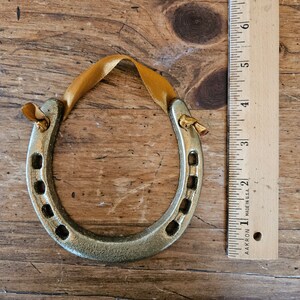 May include: A gold-colored horseshoe ornament with a gold ribbon for hanging. The horseshoe has a textured surface and is approximately 4 inches long. The ornament is displayed on a wooden surface.
