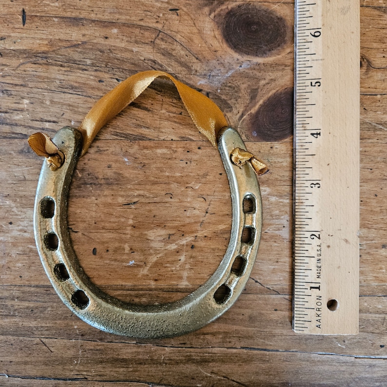 May include: A gold-colored horseshoe ornament with a gold ribbon for hanging. The horseshoe has a textured surface and is approximately 4 inches long. The ornament is displayed on a wooden surface.