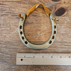 May include: A gold-colored horseshoe ornament with a yellow ribbon for hanging. The horseshoe has a distressed finish and is approximately 4 inches long. The ornament is displayed on a wooden surface.