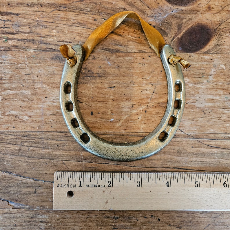 May include: A gold-colored horseshoe ornament with a yellow ribbon for hanging. The horseshoe has a distressed finish and is approximately 4 inches long. The ornament is displayed on a wooden surface.