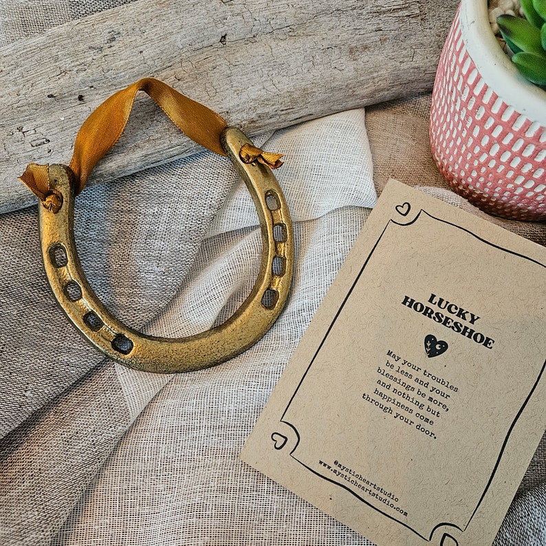 May include: A golden horseshoe with a mustard yellow ribbon, displayed on a textured surface. A card with the text "LUCKY HORSESHOE" and a poem is next to the horseshoe. The horseshoe is a symbol of good luck.