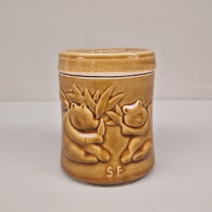 Può includere: Un barattolo di ceramica marrone con coperchio. Il barattolo presenta un disegno a rilievo di due orsi e fogliame. Le lettere "SF" sono in basso sul barattolo. Il barattolo è cilindrico e ha un coperchio abbinato.