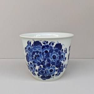 Op de afbeelding: Witte keramische bloempot met een levendig blauw bloemmotief. De bloemen zijn dicht op elkaar gepakt en vormen een dicht patroon rond de buitenkant van de pot. De rand is effen wit.