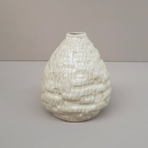 May include: A small, white ceramic vase with a textured, bumpy surface. The vase has a rounded, bulbous shape.
