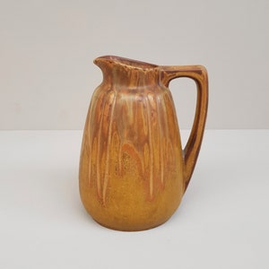 May include: A brown and yellow ceramic pitcher with a textured surface and a handle. The pitcher has a rounded body and a slightly flared lip.