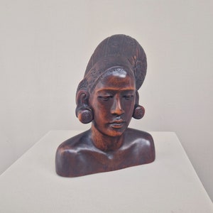 May include: A wooden sculpture of a woman's bust with a detailed hairstyle and large earrings. The sculpture is carved from dark wood and has a natural finish.