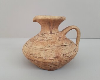 Könnte beinhalten: Eine kleine, braune Tonkrug mit rundem Körper und einem kurzen, gebogenen Henkel. Der Krug hat eine strukturierte Oberfläche und scheint handgefertigt zu sein.