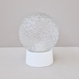 Puede incluir: Una esfera de vidrio transparente con una superficie texturizada con forma de burbujas se asienta sobre una base blanca cilíndrica. La esfera es redonda y parece estar hecha de vidrio. La base es blanca, opaca y sólida. El fondo es claro.