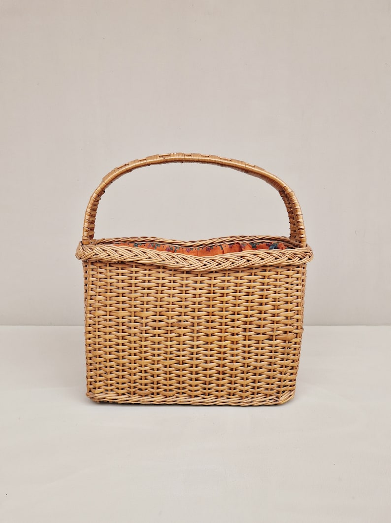 May include: A woven wicker basket with a single handle. The basket has a rectangular shape and a brown, natural color. The inside of the basket is lined with a floral patterned fabric.