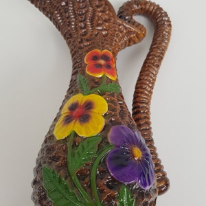 May include: Brown ceramic pitcher with a woven basket texture, adorned with colorful pansy flowers. The pitcher has a curved handle and a spout. The flowers are yellow, purple, and red with green leaves, adding a touch of color to the brown pitcher.