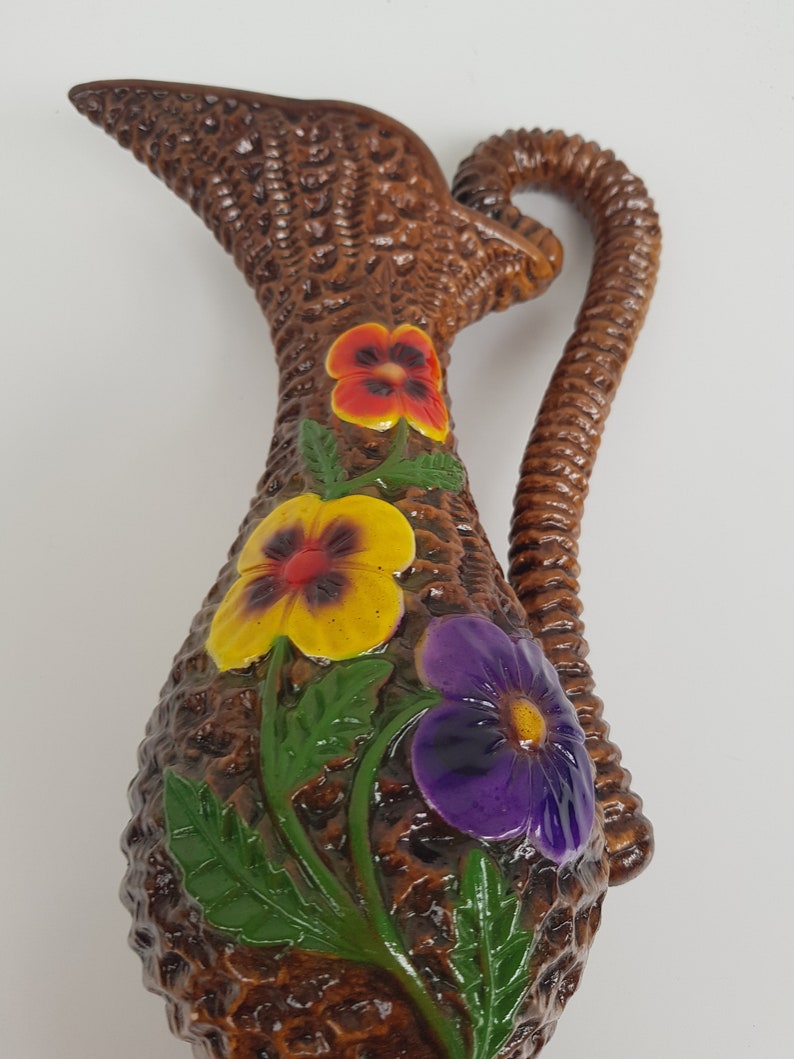 May include: Brown ceramic pitcher with a woven basket texture, adorned with colorful pansy flowers. The pitcher has a curved handle and a spout. The flowers are yellow, purple, and red with green leaves, adding a touch of color to the brown pitcher.