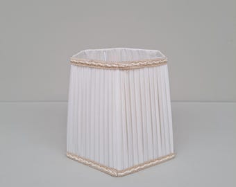Könnte beinhalten: Ein weißer, plissierter Lampenschirm mit quadratischer Form. Der Schirm ist oben und unten mit einer beigefarbenen, geflochtenen Bordüre versehen. Der Lampenschirm ist ein klassisches Design, das für verschiedene Einrichtungsstile geeignet ist.