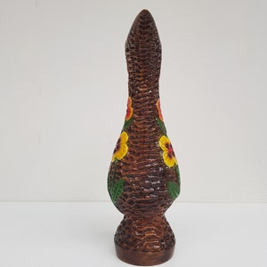 May include: A tall, brown ceramic vase with a textured, woven appearance. The vase is adorned with yellow and red floral designs and green leaves. The vase has a flared base and a long, slender neck, and is approximately 12 inches tall.