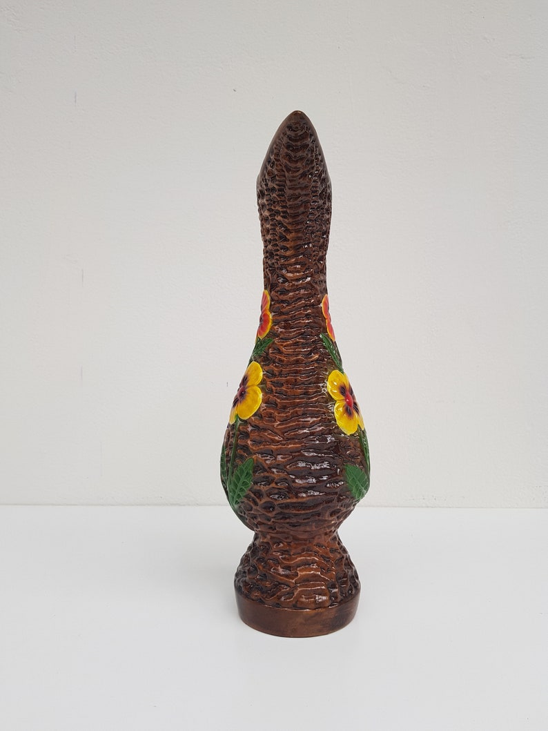 May include: A tall, brown ceramic vase with a textured, woven appearance. The vase is adorned with yellow and red floral designs and green leaves. The vase has a flared base and a long, slender neck, and is approximately 12 inches tall.