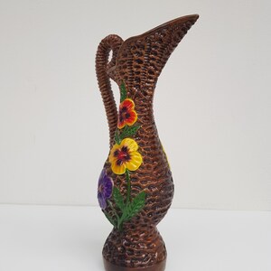 May include: Brown ceramic pitcher with a woven basket design. The pitcher features a handle and spout, and is decorated with colorful pansy flowers in red, yellow, and purple. The base is a darker brown.