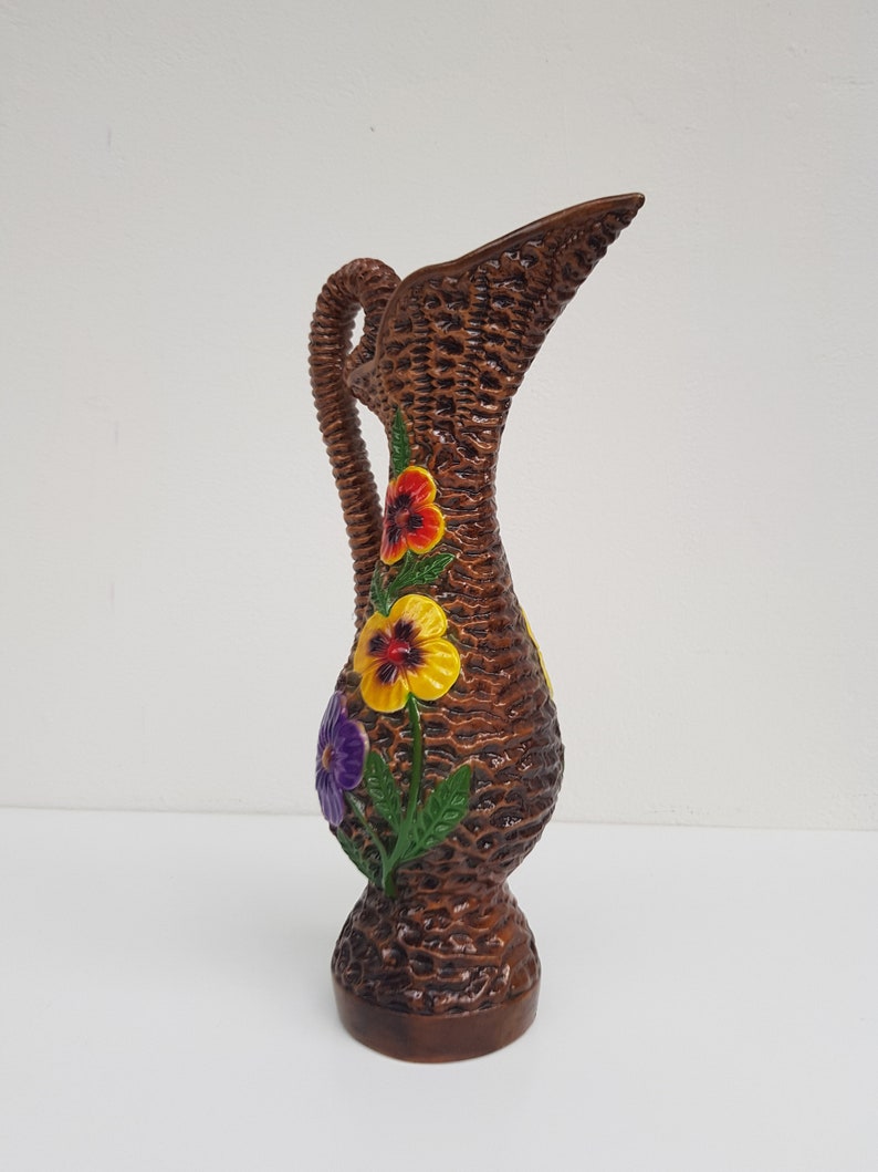 May include: Brown ceramic pitcher with a woven basket design. The pitcher features a handle and spout, and is decorated with colorful pansy flowers in red, yellow, and purple. The base is a darker brown.