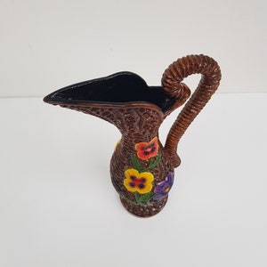 May include: A brown wicker pitcher with a woven handle and a dark interior. The pitcher is decorated with colorful pansy flowers in red, yellow, and purple. The pitcher is a decorative item.