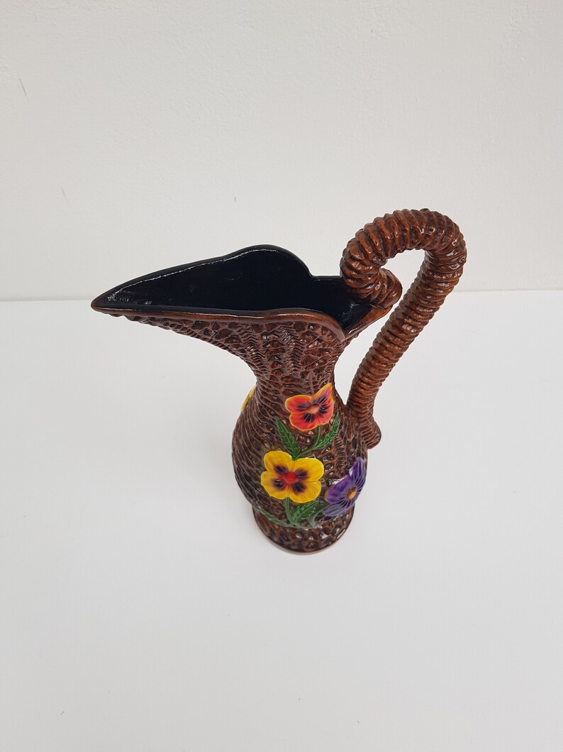May include: A brown wicker pitcher with a woven handle and a dark interior. The pitcher is decorated with colorful pansy flowers in red, yellow, and purple. The pitcher is a decorative item.