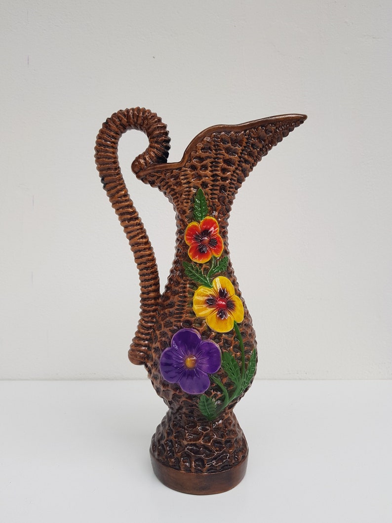 May include: A brown decorative pitcher with a woven basket design. The pitcher features a curved handle and is adorned with three colorful flower accents: red, yellow, and purple. The pitcher is approximately 10 inches tall.