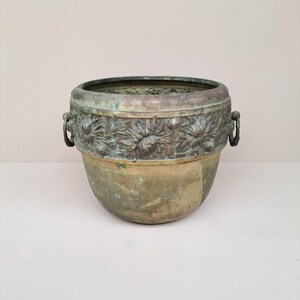 Könnte beinhalten: Ein runder, verwitterter Pflanzkübel aus Bronze mit einem markanten Reliefband aus skulpturalen Blumenmotiven am oberen Rand. Das Gefäß weist eine matte, oxidierte grün-braune Patina auf und hat zwei kleine, runde Metallringgriffe an den Seiten.