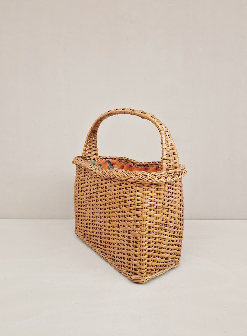 May include: A woven wicker basket with a single handle. The basket is rectangular with a slightly rounded top and a natural brown color.