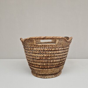 May include: A large, round, woven wicker basket with two handles. The basket is made of natural brown wicker and has a slightly flared shape.