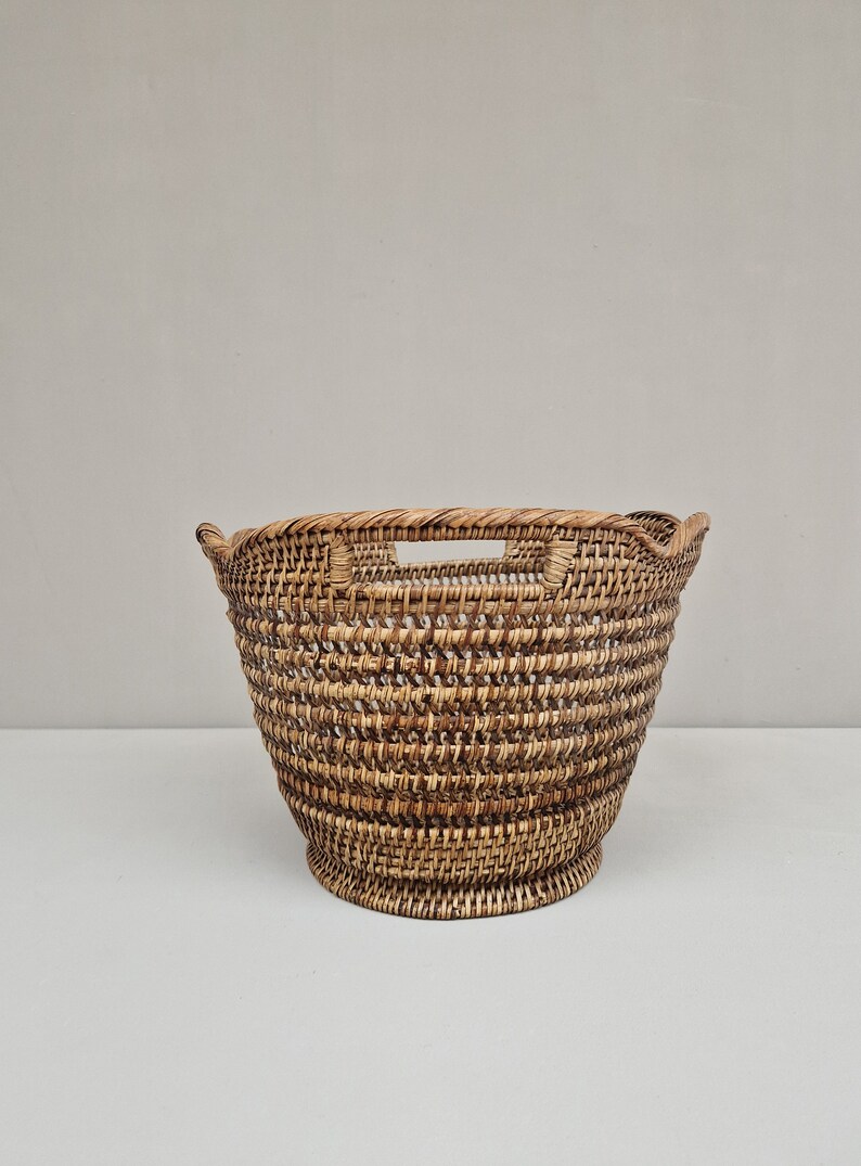 May include: A large, round, woven wicker basket with two handles. The basket is made of natural brown wicker and has a slightly flared shape.