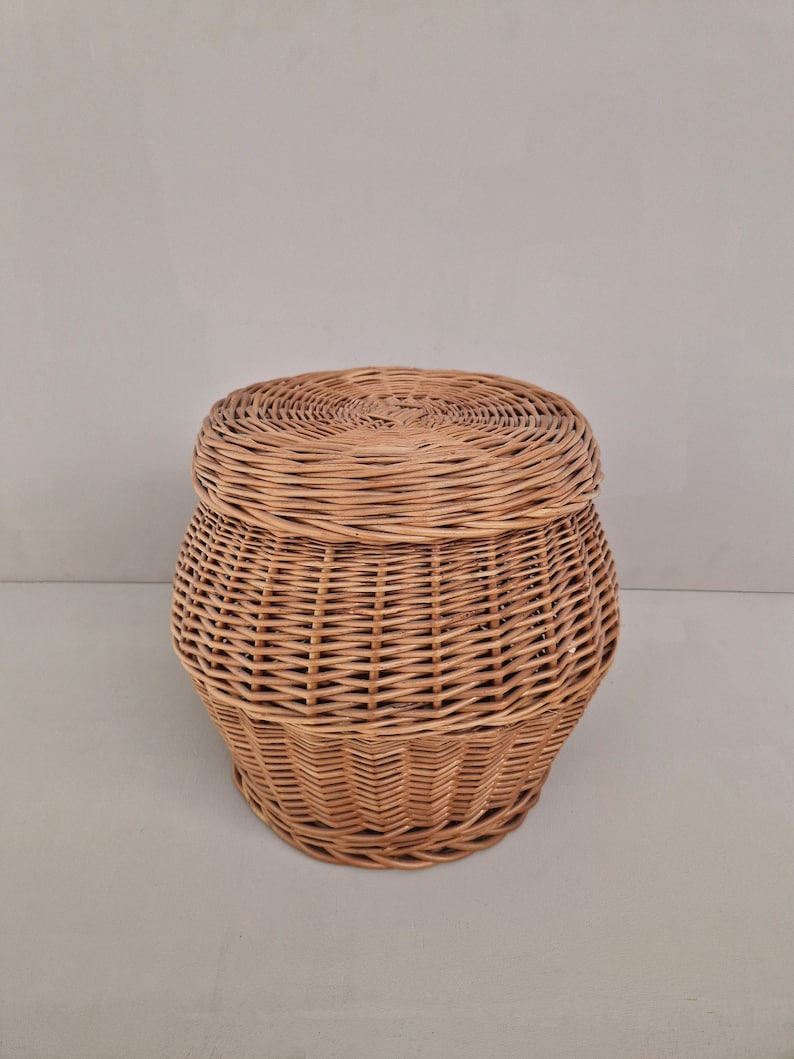 May include: A round, woven wicker basket with a lid. The basket is light brown and has a natural, rustic look.