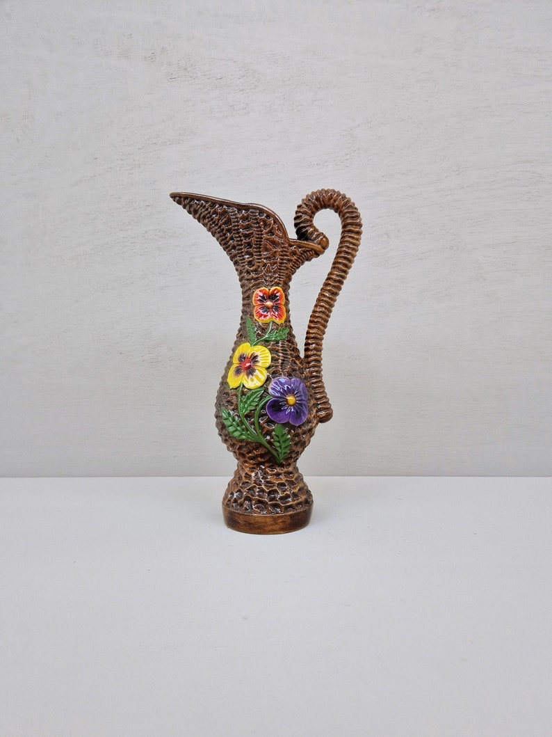 May include: A brown ceramic pitcher with a woven basket design. The pitcher features a curved handle and is adorned with three colorful pansy flower appliques. The flowers are red, yellow, and purple.