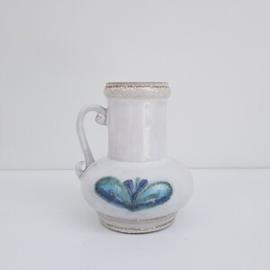 K&ouml;nnte beinhalten: Eine wei&szlig;e Keramikvase mit einem blauen und gr&uuml;nen Blumendesign. Die Vase hat einen runden K&ouml;rper und einen kurzen Hals mit einem Henkel.