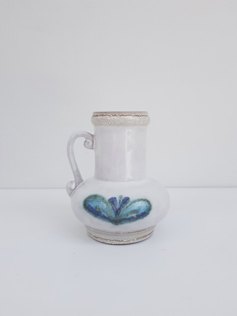 K&ouml;nnte beinhalten: Eine wei&szlig;e Keramikvase mit einem blauen und gr&uuml;nen Blumendesign. Die Vase hat einen runden K&ouml;rper und einen kurzen Hals mit einem Henkel.