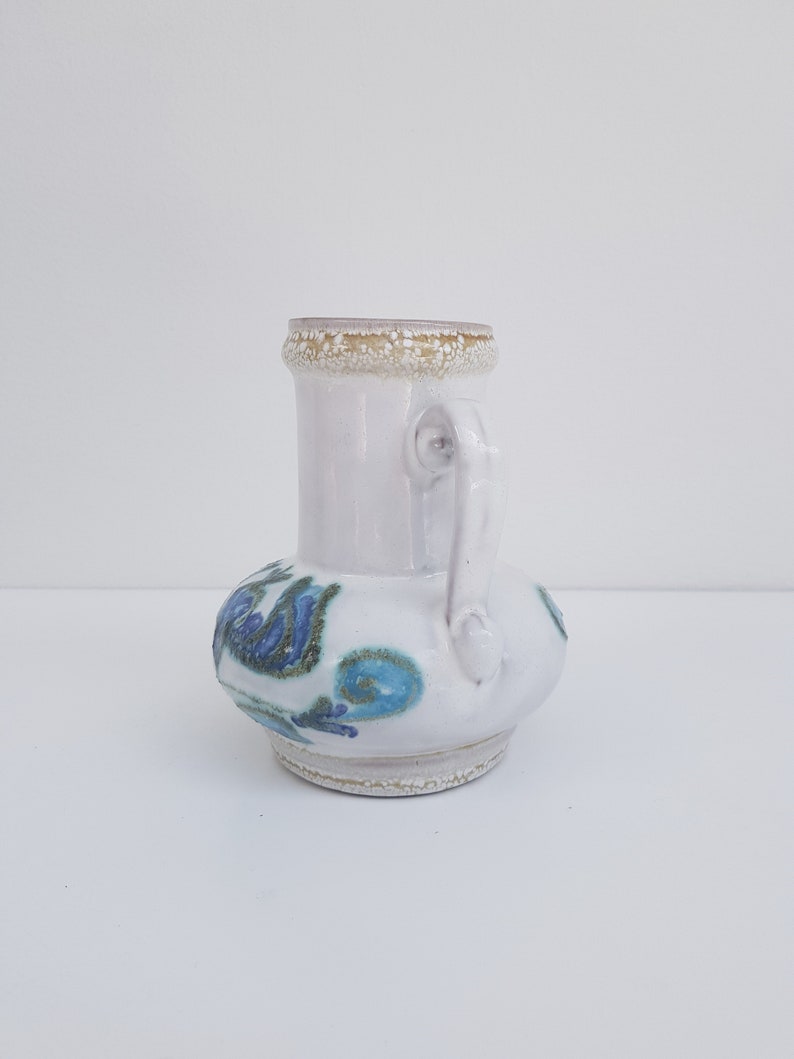 K&ouml;nnte beinhalten: Wei&szlig;e Keramikvase mit blauem und gr&uuml;nem Blumendesign. Die Vase hat einen runden K&ouml;rper und einen kleinen Henkel.