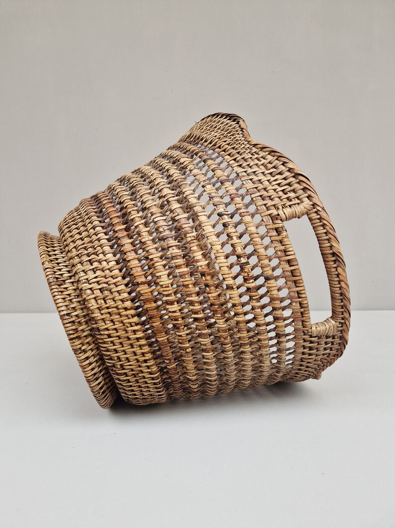 May include: A woven wicker basket with a rounded shape and a handle on the side. The basket is made of natural brown rattan.